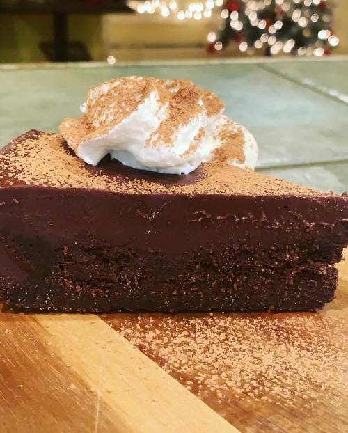 A slice of rich chocolate cake topped with whipped cream and cocoa powder, resting on a wooden surface.