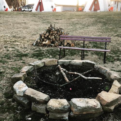 A stone fire pit with sticks inside, a bench nearby, and a stack of firewood in the background with teepee structures.
