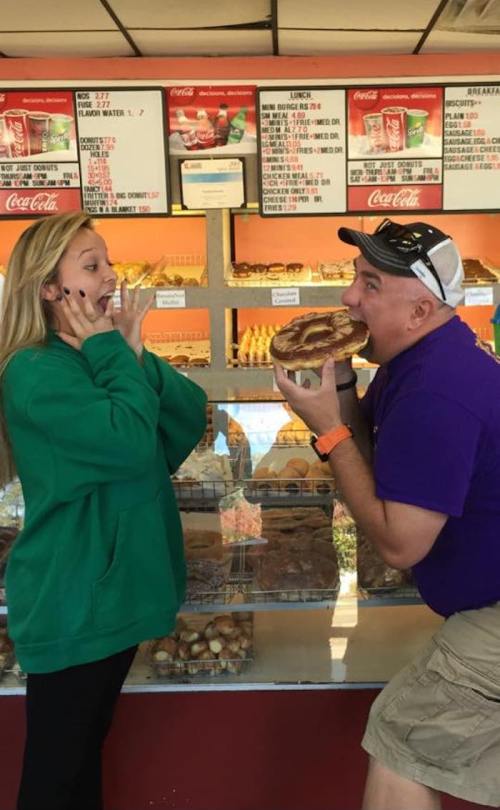 A man and woman playfully pose in front of a bakery display, with the man pretending to eat a large pastry.