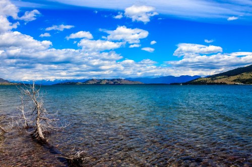 A serene lake with clear blue water, surrounded by mountains and a bright sky filled with fluffy clouds.