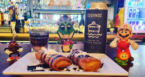 A colorful bar scene featuring a can of beer, a shot glass, and two dessert pastries drizzled with chocolate and powdered sugar.