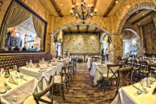 Elegant restaurant interior with wooden floors, stone walls, and tables set for dining under a chandelier.