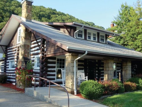A charming log cabin-style house with a stone chimney, rocking chairs on the porch, and lush greenery surrounding it.