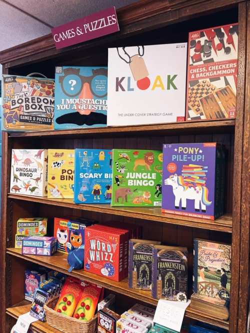 A wooden shelf displaying various board games and puzzles, including "Klook," "Scary Bin," and "Pony-Pile-Up."
