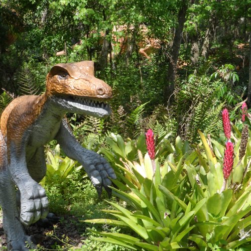 A dinosaur statue in a lush garden, surrounded by vibrant green plants and pink flowers.