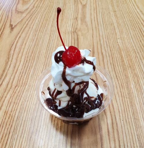 A dessert cup with swirls of whipped cream, chocolate sauce, and a cherry on top, placed on a wooden table.