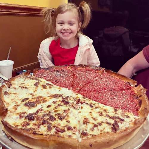 A smiling girl in a red shirt sits beside a large pizza with half cheese and half deep-dish toppings.