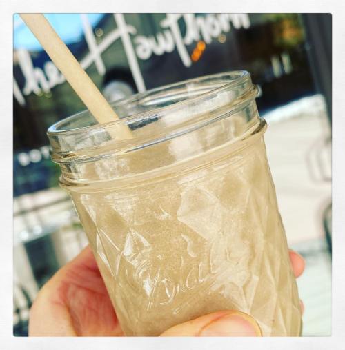 A hand holding a mason jar filled with a frosty beverage and a wooden straw, with a blurred background.
