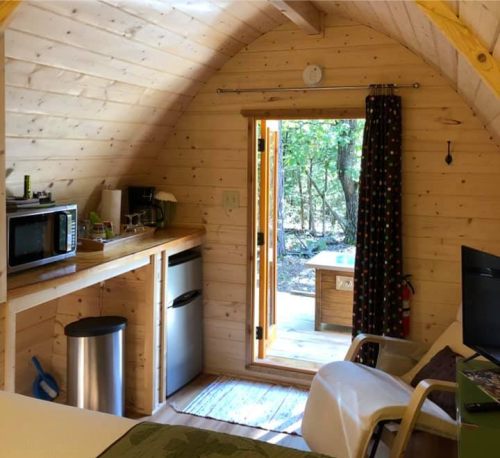 Cozy wooden cabin interior with a kitchenette, door to the outdoors, and a small table visible outside.