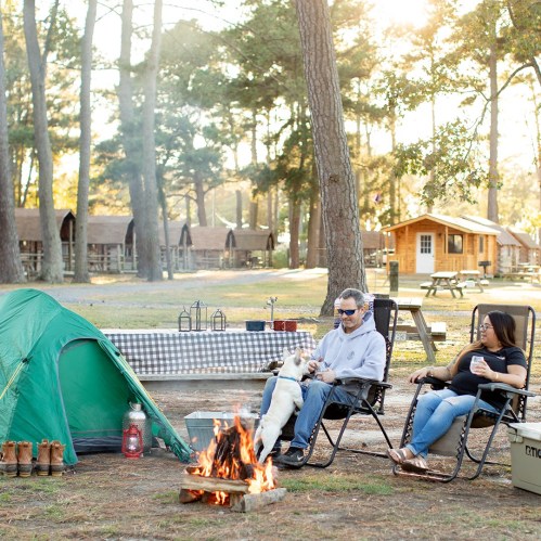 A couple relaxes by a campfire with a dog, surrounded by tents and cabins in a wooded campsite.