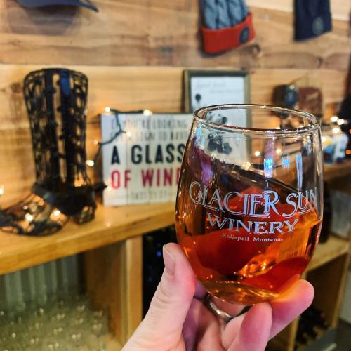 A hand holding a glass of wine with "Glacier Sun Winery" etched on it, set against a rustic wooden backdrop.
