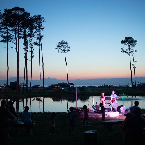 A band performs by a lake at sunset, with trees and tents in the background and an audience enjoying the music.