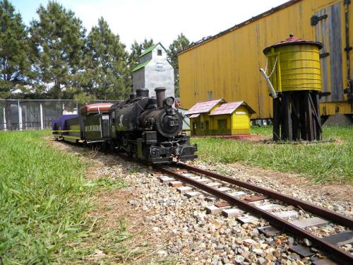 A miniature steam train on tracks, surrounded by small buildings and greenery, with a yellow water tower in the background.