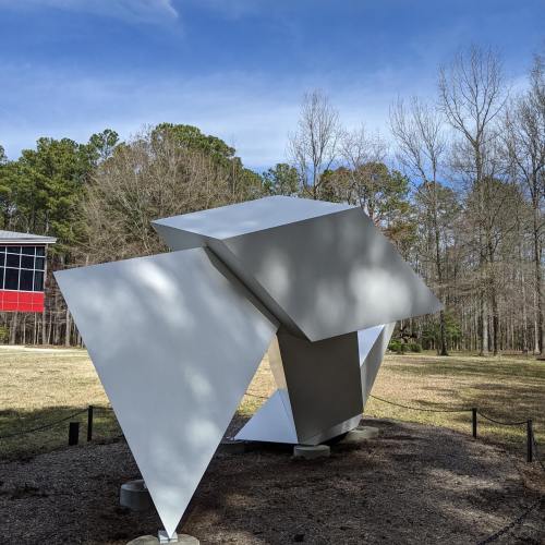Abstract white sculpture with angular shapes, set in a grassy area surrounded by trees and a building in the background.