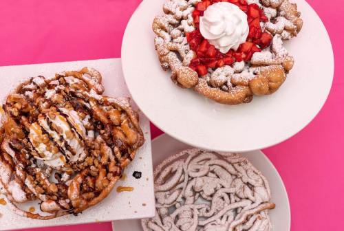 Three plates of funnel cakes topped with whipped cream, strawberries, and drizzled chocolate, set against a pink background.