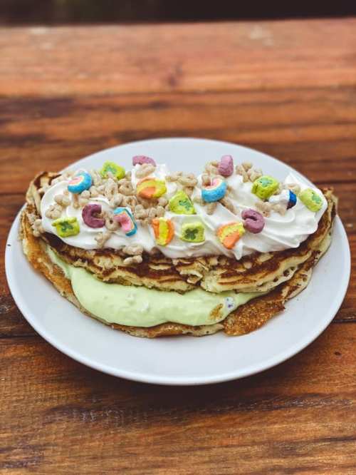 A stack of pancakes topped with whipped cream, colorful cereal, and a green cream on a white plate.