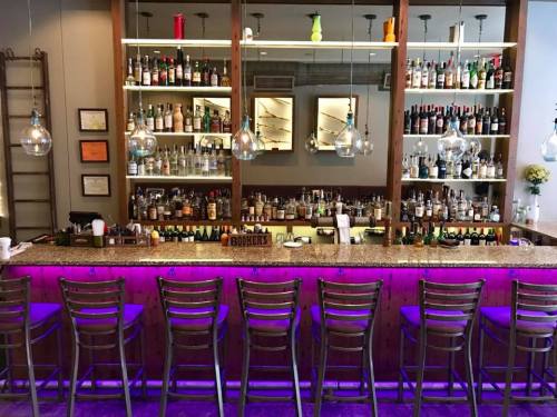 A stylish bar with a well-stocked shelf of bottles, illuminated by pendant lights and featuring a purple-lit counter.