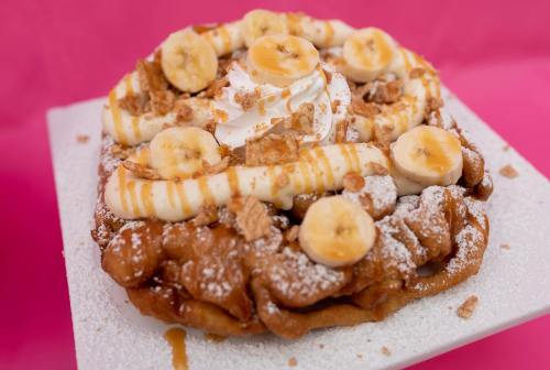A delicious funnel cake topped with banana slices, whipped cream, and crushed cookies, dusted with powdered sugar.