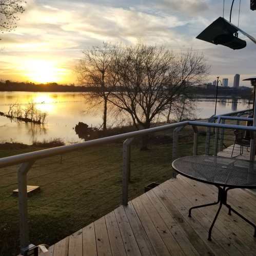 A serene sunset over a lake, with trees and a deck in the foreground, reflecting warm colors in the water.