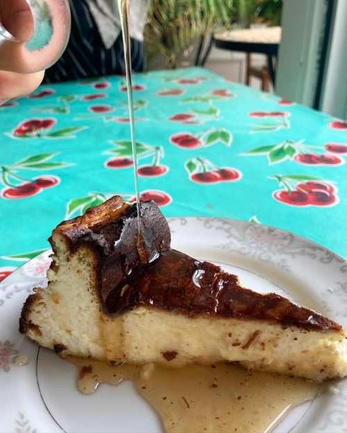 A slice of cheesecake drizzled with syrup on a decorative plate, set on a cherry-patterned tablecloth.