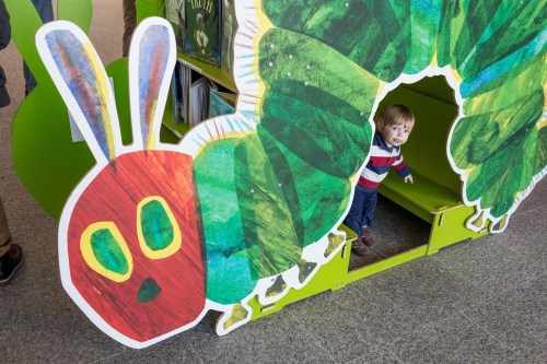 A child peeks out from a colorful caterpillar-shaped play structure in a library.