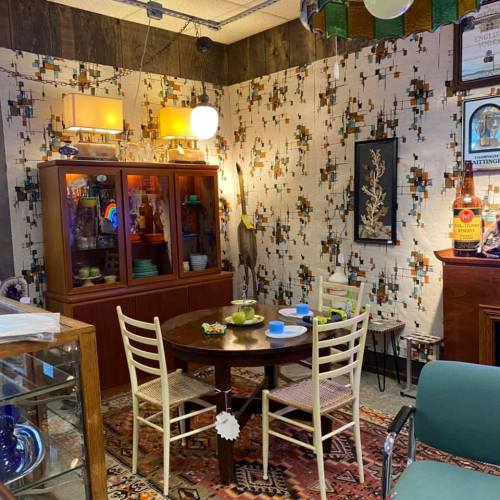 A cozy vintage dining area with a round table, four chairs, a cabinet, and patterned wallpaper.