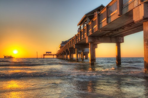 A sunset view of a pier extending over calm waters, with warm orange and yellow hues illuminating the sky.