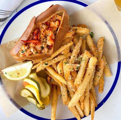 A lobster roll in a toasted bun served with crispy fries and lemon wedges on a white plate.