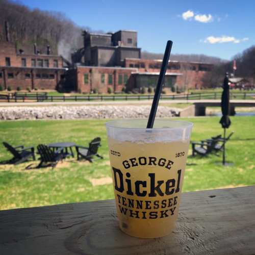 A cup of George Dickel Tennessee Whiskey sits on a table, with a scenic view of a distillery and green landscape in the background.