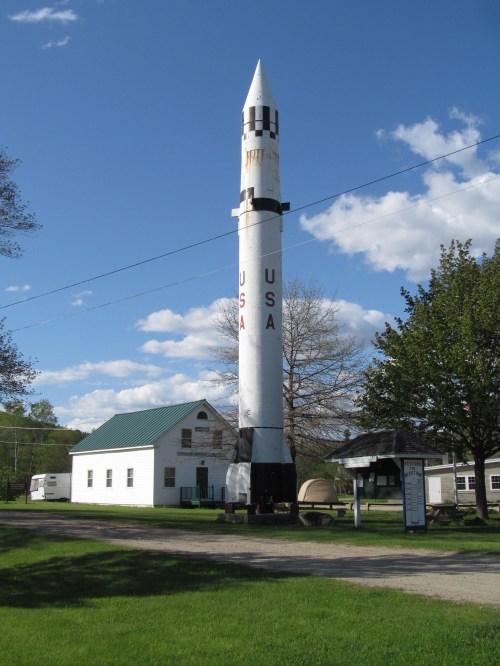 A tall rocket with "USA" written on it stands beside a small white building under a blue sky with fluffy clouds.
