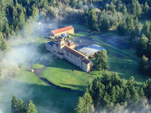 Aerial view of a large building surrounded by trees and mist, with additional structures and a parking area nearby.