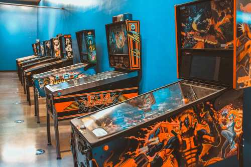 A row of colorful pinball machines against a blue wall, featuring various themes and designs.