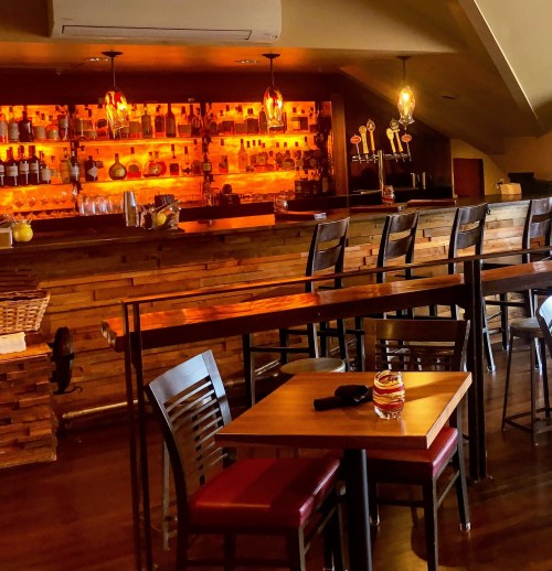 A cozy bar with wooden decor, illuminated shelves of bottles, and empty tables and chairs.
