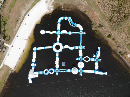 Aerial view of a large inflatable water park on a lake, featuring various slides and obstacles in blue and white.