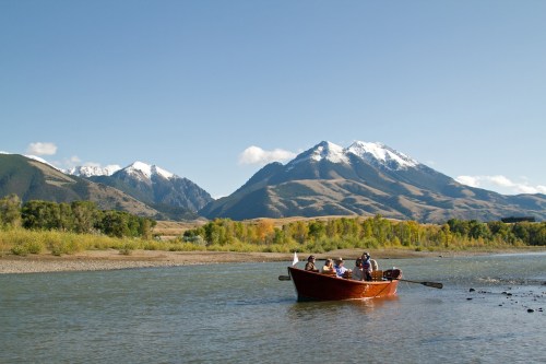 A wooden boat with people rowing on a river, surrounded by mountains and clear blue skies.