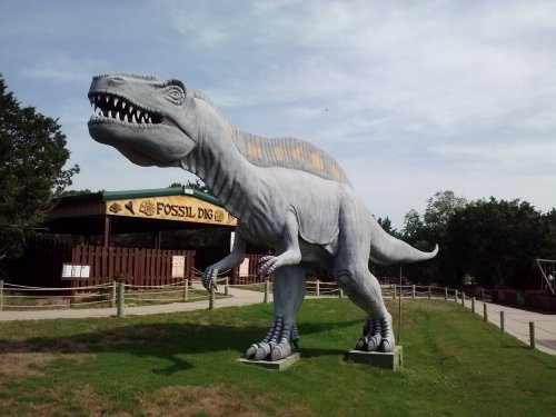 A large dinosaur statue stands outside a building labeled "Fossil Dig," surrounded by grass and trees.