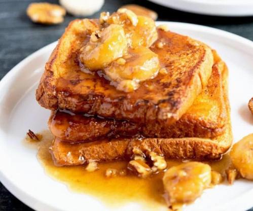 Stack of French toast topped with caramelized bananas and nuts, drizzled with syrup on a white plate.