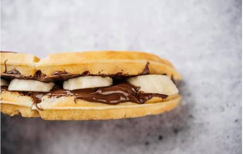 A waffle sandwich filled with banana slices and chocolate spread, set against a textured background.