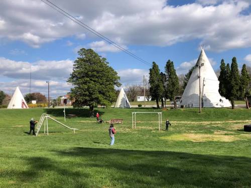 A grassy park with teepee structures, playground equipment, and people enjoying the outdoors under a partly cloudy sky.