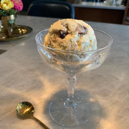 A scoop of ice cream with chocolate chunks in a glass dish, accompanied by a gold spoon on a marble surface.