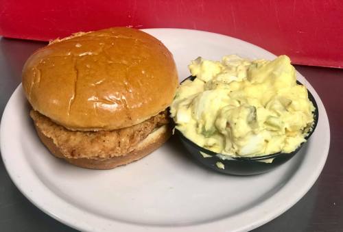 A plate with a fried chicken sandwich on a bun and a side of creamy potato salad in a small bowl.
