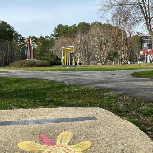 A colorful outdoor sculpture stands among trees, with a pathway and a stone marker featuring a flower design in the foreground.