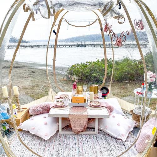 A cozy outdoor picnic setup inside a clear dome, featuring pillows, a table with drinks, and a scenic waterfront view.