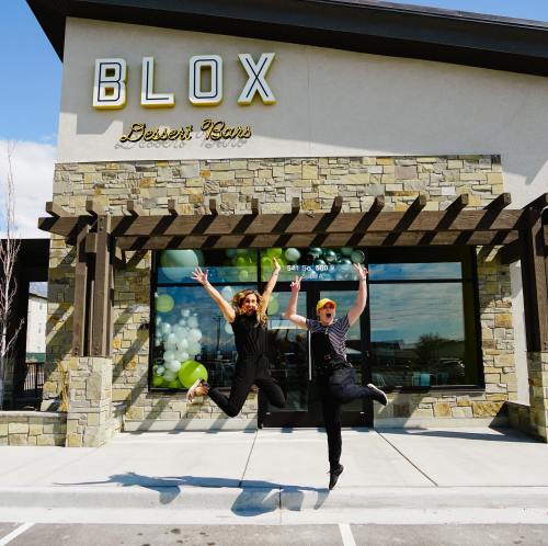 Two people joyfully jumping in front of a dessert shop called "BLOX," decorated with balloons and a stone facade.
