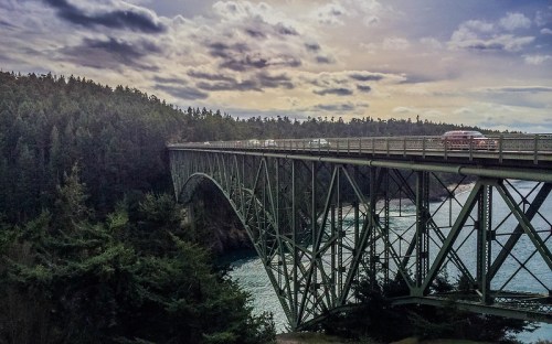 A steel bridge spans a river, surrounded by lush green trees and a cloudy sky.