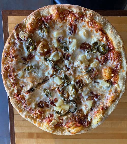 A round pizza topped with cheese, pineapple, jalapeños, and pieces of bacon on a wooden board.