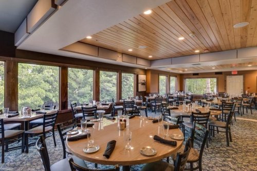 A spacious restaurant interior with wooden ceilings, round tables set for dining, and large windows overlooking greenery.