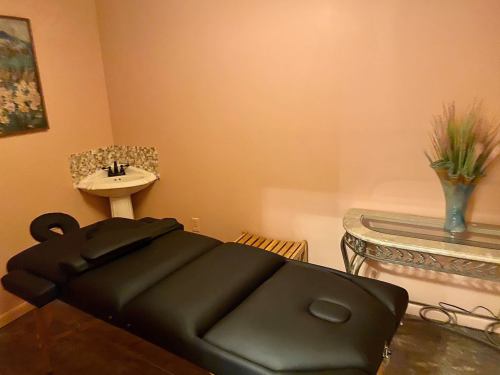 A massage room featuring a black massage table, a small sink, and a decorative vase on a table against a pink wall.