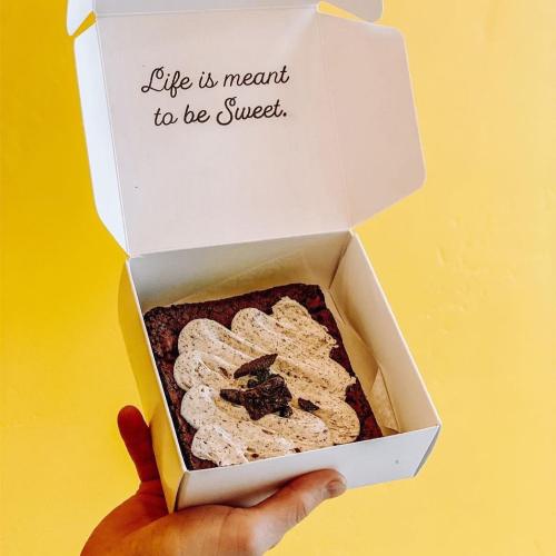 A hand holds a white box with a brownie topped with cream, and the message "Life is meant to be Sweet" inside.