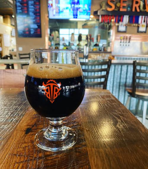 A glass of dark beer with a logo sits on a wooden table, with a bar and TV in the background.